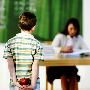 Boy (8-9) holding apple behind back, looking at teacher in classroom, focus on boy